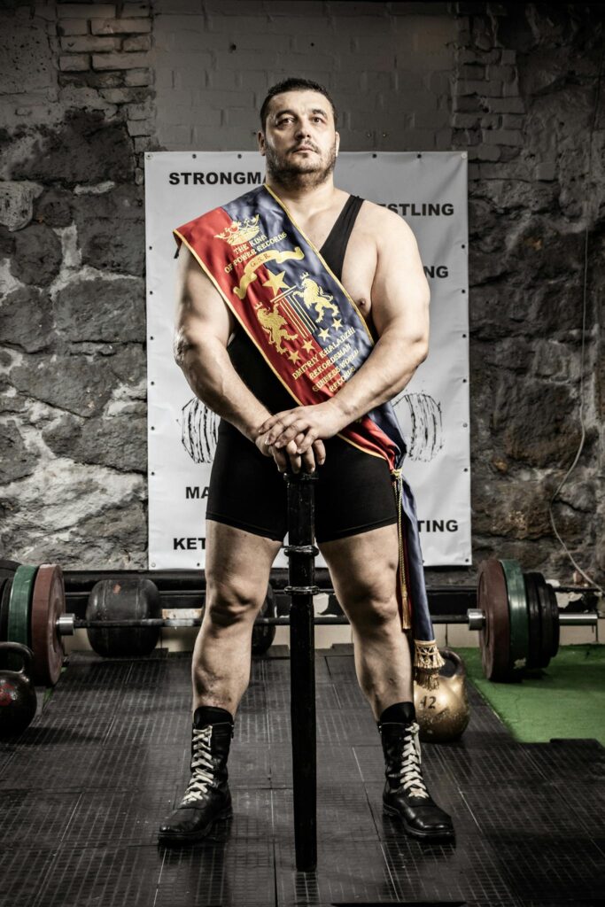 Muscular strongman wearing a championship sash stands confidently in a gym holding a kettlebell.