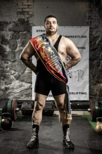 Muscular wrestler with champion banner stands confidently in gym setting.